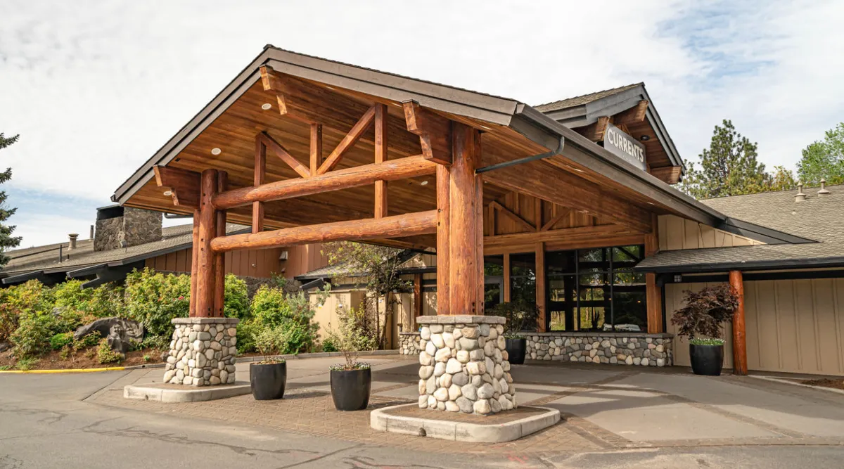 This exterior view focuses on the glass entrance framed by large wooden beams and stone pillars, surrounded by well-kept greenery. The image suggests a welcoming, accessible property with ample parking and a practical layout for guests arriving with gear or pets.