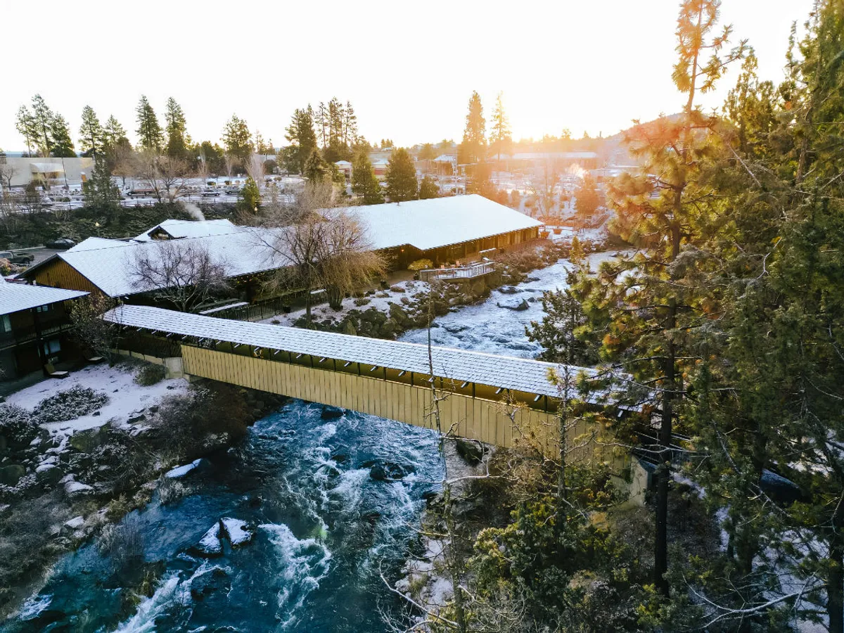 This image captures a covered bridge spanning the Deschutes River, with snow-covered roofs and pine trees creating a tranquil, natural setting. The river runs through the scene, inviting exploration and outdoor activities, while the bridge provides a direct connection to walking trails along the riverbank.