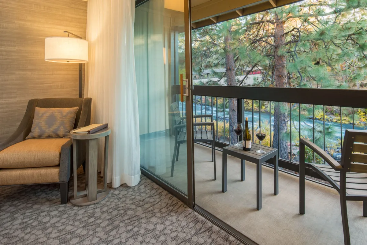A tranquil corner of the superior room with a comfortable armchair, a small side table, and a glass sliding door that opens to a private balcony. Wine glasses on the table suggest relaxation, and the balcony offers a scenic view, capturing the riverside ambiance mentioned in the section.