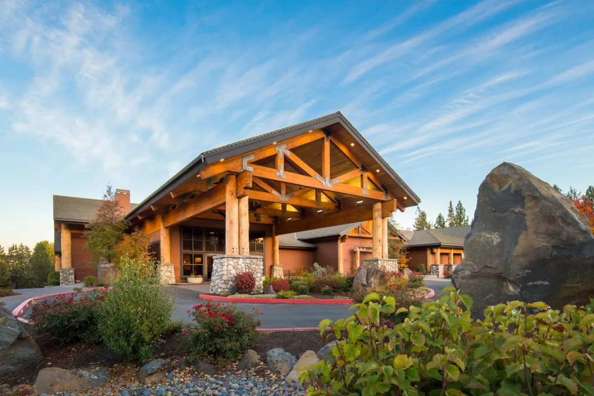 This exterior image highlights the property’s inviting entrance, featuring large wooden beams, a stone façade, and lush landscaped gardens. The scene suggests easy transition from indoor comfort to outdoor adventure, with paths and greenery visible that hint at nearby trails. The welcoming atmosphere aligns with a sense of arrival ready for exploration.