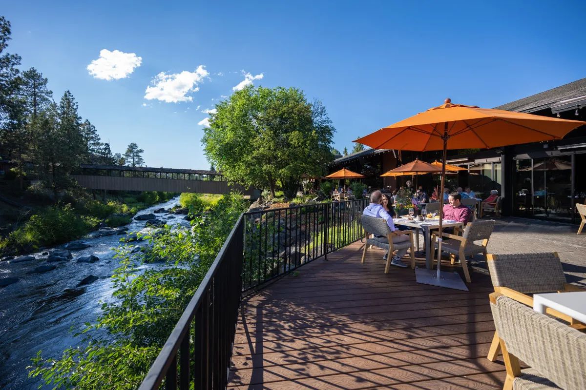 An exterior scene featuring the Deschutes River bordered by green trees, with outdoor seating shaded by orange umbrellas. Visitors relax and socialize along the riverbank, creating a lively yet natural atmosphere perfect for unwinding after a day of adventure.