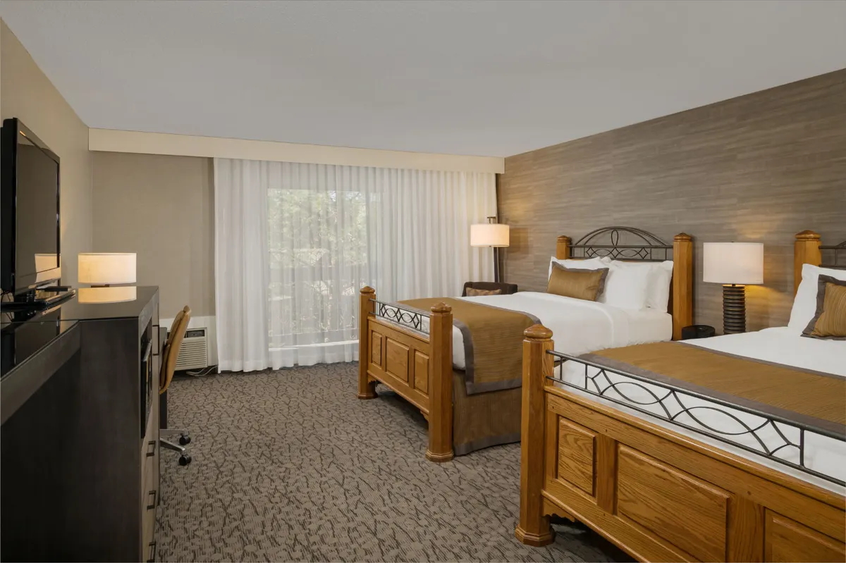 This image shows a superior room with two comfortable wooden queen beds, a large flat-screen TV, soft lighting from side lamps, and curtains that lead to a view outside. The arrangement feels spacious and inviting, perfect for relaxing after a busy day or enjoying entertainment in-room.