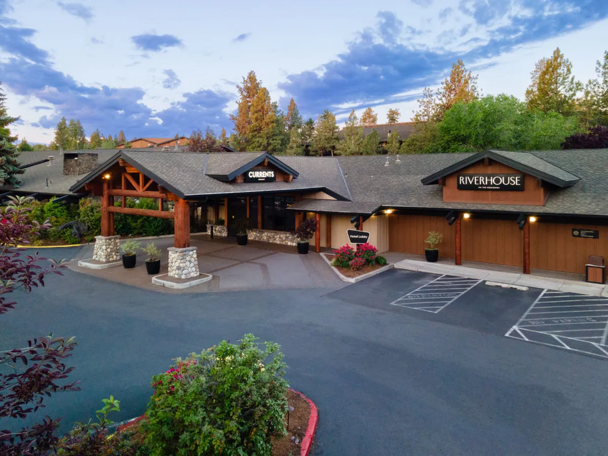 The entrance area of Riverhouse Lodge, framed by lush landscaping and clear signage for CURRANTS and RIVERHOUSE. The inviting canopy and bustling exterior set the scene for arrival and departure before and after outdoor excursions.