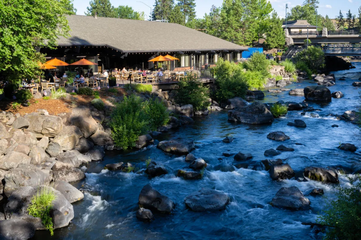 The image showcases a stunning outdoor restaurant patio perched beside the flowing Deschutes River, framed by dramatic rock formations and lush greenery. Guests are seen relaxing, sipping drinks, and socializing as the sun sets, casting a golden glow over the tranquil water and natural landscape.