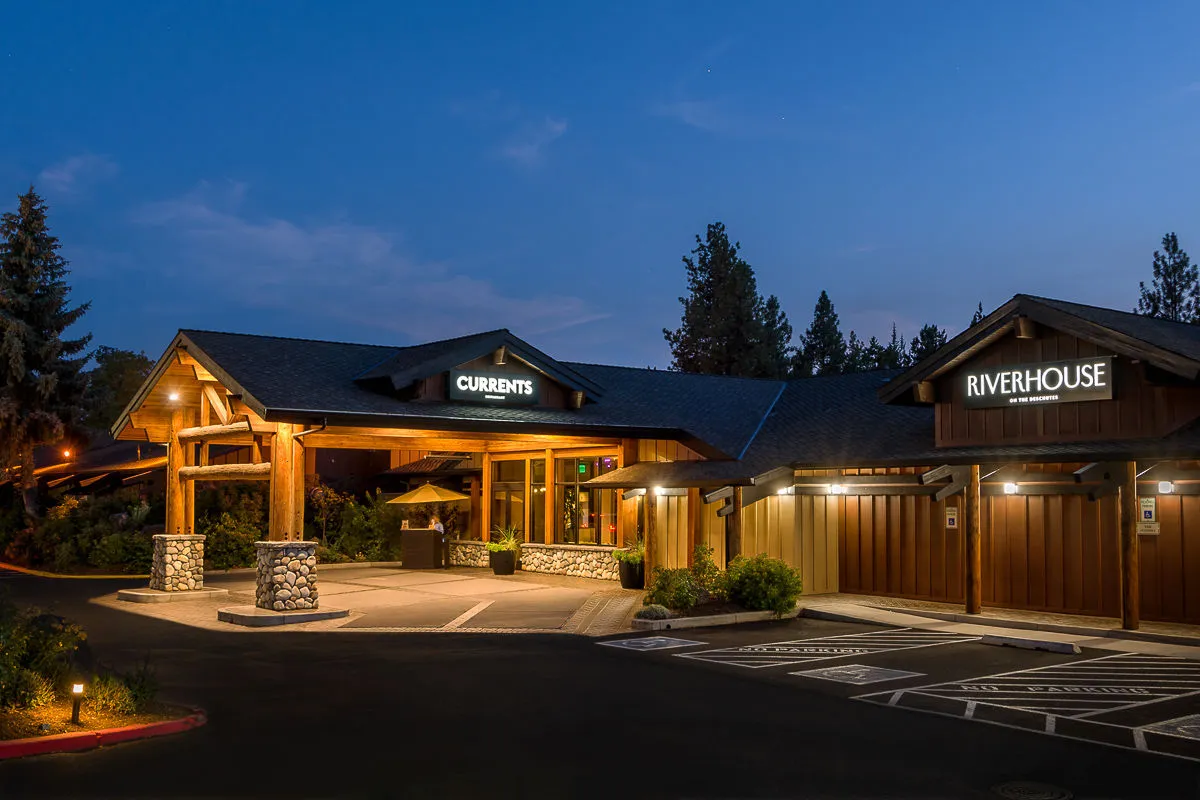 The image captures the exterior entrance to Riverhouse Lodge, featuring a welcoming awning, clear signage for the on-site dining options (CURRANTS and RIVERHOUSE), attractive landscaping, and sturdy stone columns. The view suggests a tranquil and accessible space where guests can arrive and immediately sense the lodge's inviting atmosphere. Hints of sunset and patio ambiance give a glimpse of social and family activities that can take place outdoors.