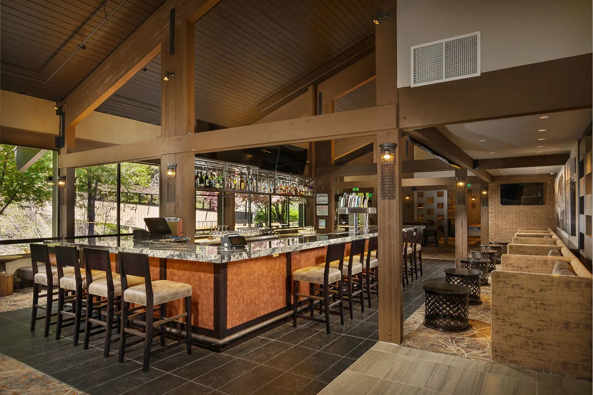 A contemporary lounge and bar area, with a sleek bar counter, comfortable bar stools, a cozy sitting area, and an attractive display of bottles. The ambiance is lively yet relaxed, ideal for socializing, drinks, and watching sports, as suggested by the presence of TVs.