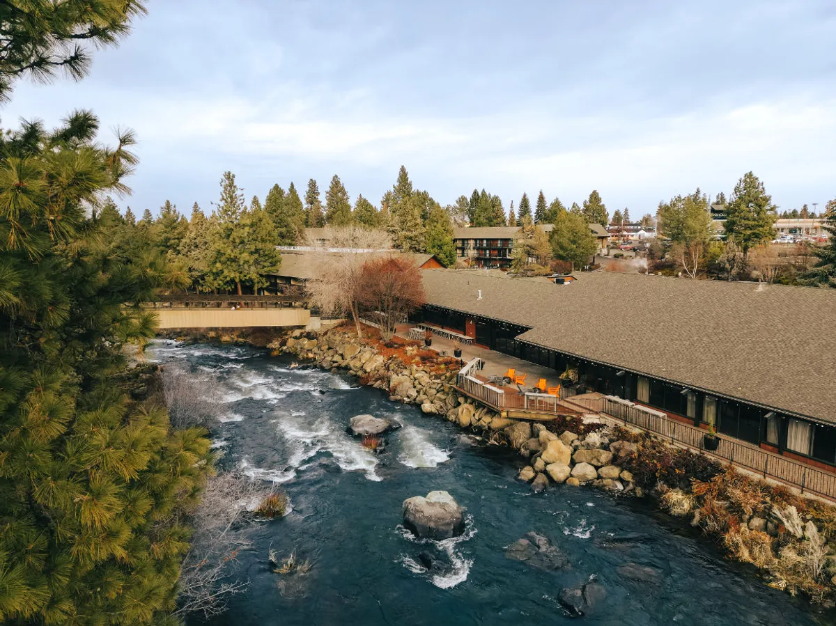 This serene exterior shot captures the river, building structure, deck area, and surrounding trees. It conveys a peaceful connection to nature, emphasizing proximity to riverside trails and opportunities for outdoor activities like nature walks and relaxation.